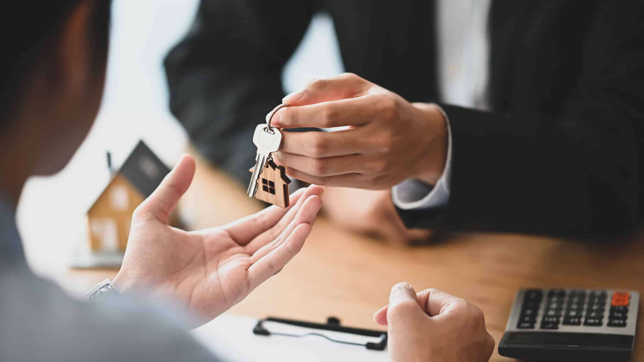 Cropped shot of House broker giving a property key to his customer while he has writing/signing on agreement at the modern wooden desk. Loan, Debt, Credit, Buying or Selling agreement concept.