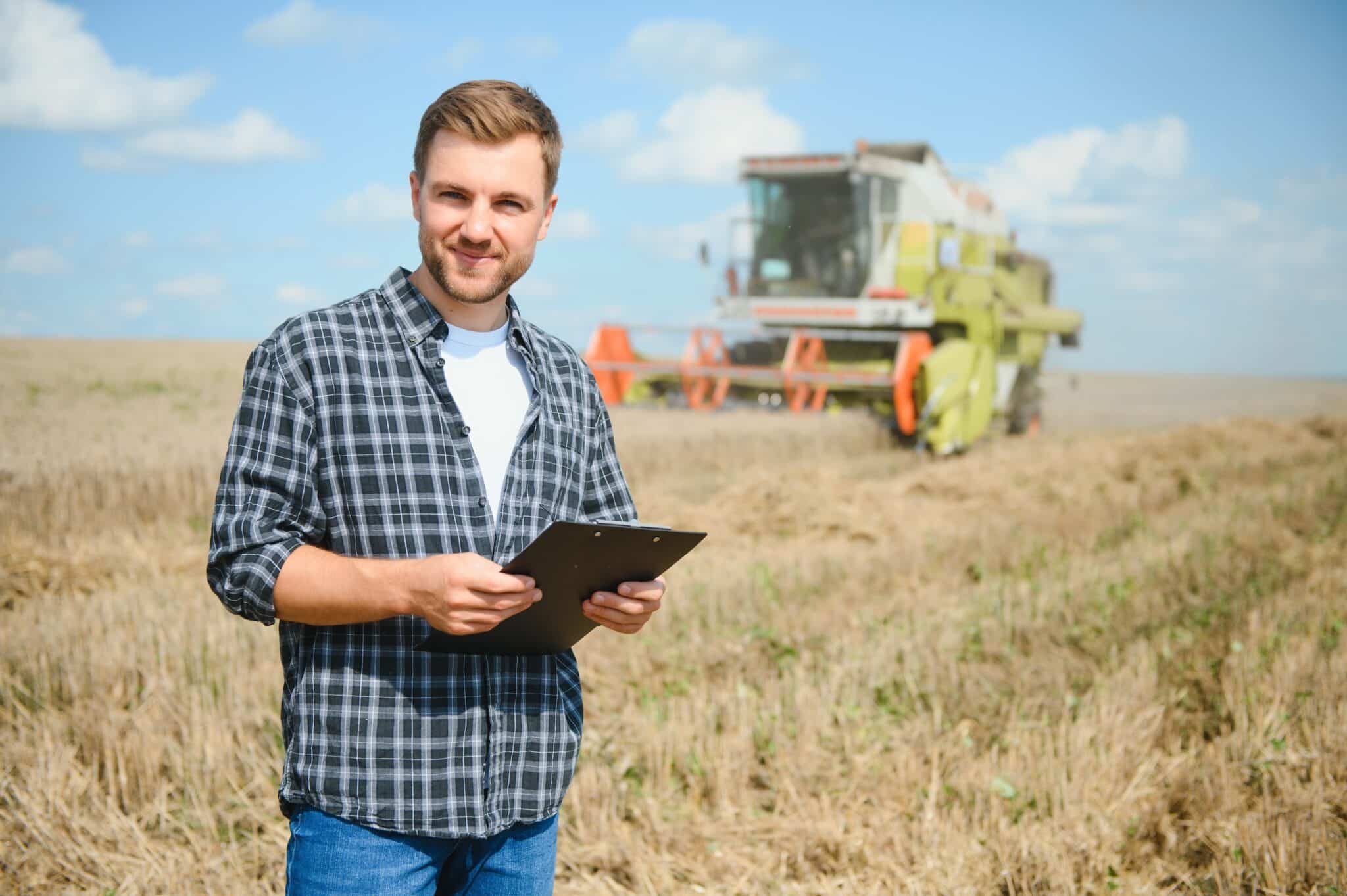 Happy farmer proudly standing in a field. Combine harvester driver going to crop rich wheat harvest. Agronomist wearing flannel shirt, looking at camera on a farmland.